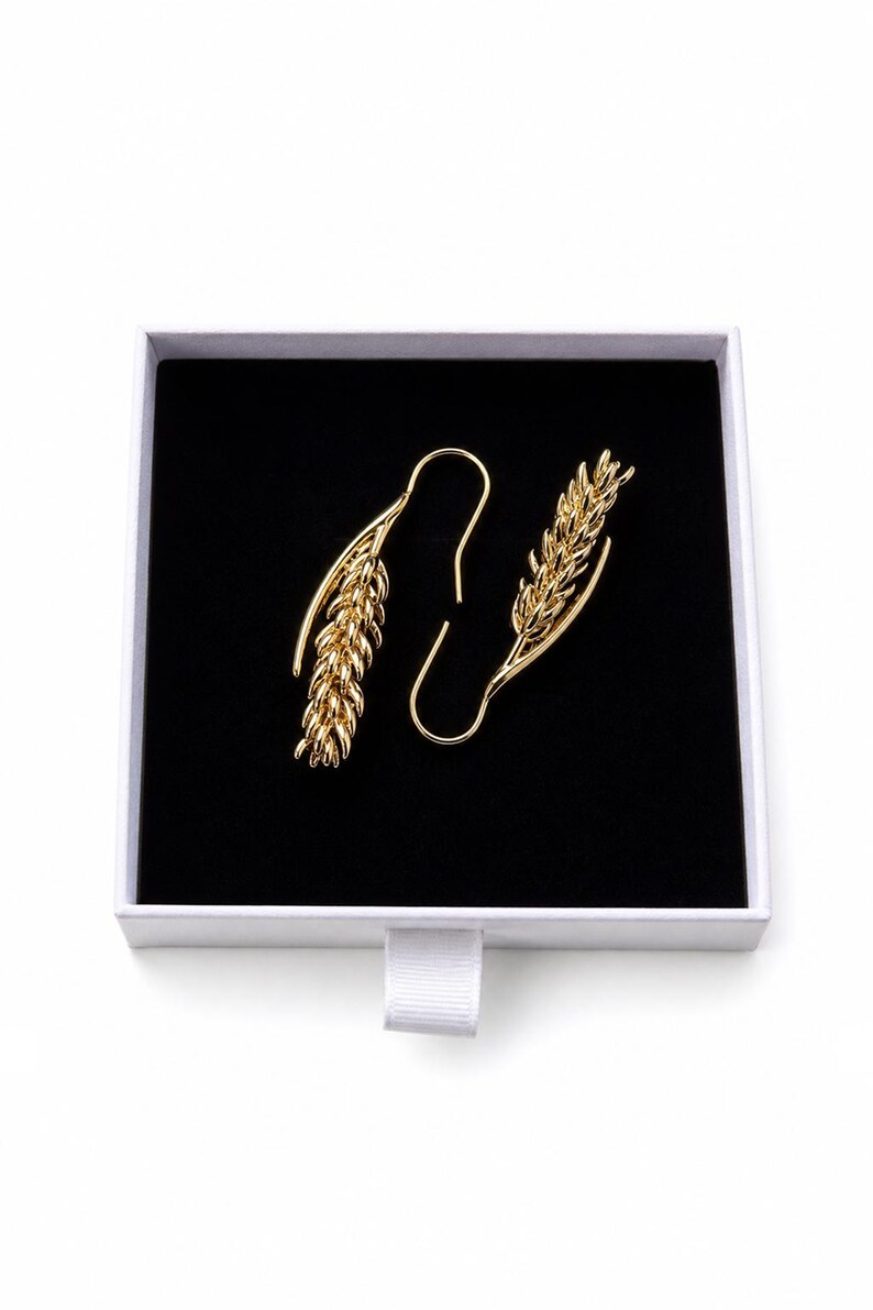 May include: A pair of gold-colored earrings shaped like wheat stalks, presented in a white box with a black interior. The earrings have a hook design for pierced ears and are displayed against a contrasting black background.