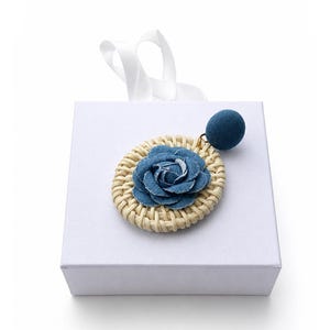 May include: A single earring featuring a circular woven rattan base topped with a blue denim fabric rose. A small, round, blue fabric-covered stud is attached to the top of the rattan circle by a gold-toned metal jump ring, resting on a white box.