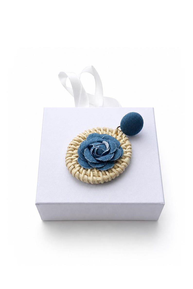 May include: A single earring featuring a circular woven rattan base topped with a blue denim fabric rose. A small, round, blue fabric-covered stud is attached to the top of the rattan circle by a gold-toned metal jump ring, resting on a white box.