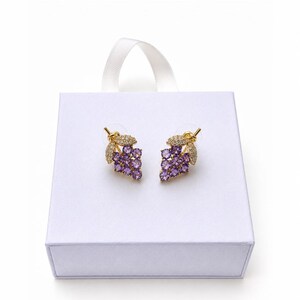 K&ouml;nnte beinhalten: Ein Paar goldfarbene Ohrstecker in Form von Trauben, jeweils mit kleinen lila Steinen. Die Ohrringe werden auf einer wei&szlig;en Schachtel mit wei&szlig;em Schleifenband pr&auml;sentiert.