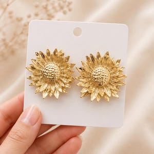 May include: A pair of gold-coloured sunflower earrings. Each earring features a detailed sunflower design with textured petals and a central disc. The earrings are displayed on a white card, showcasing the jewellery's intricate details.