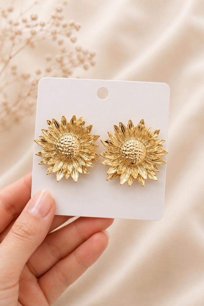 May include: A pair of gold-coloured sunflower earrings. Each earring features a detailed sunflower design with textured petals and a central disc. The earrings are displayed on a white card, showcasing the jewellery's intricate details.