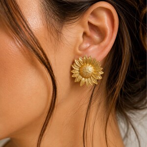 May include: Gold-coloured sunflower earrings. The earrings feature detailed petals and a textured centre, resembling a real sunflower. The earrings are worn on an ear, with the person's skin visible in the background.