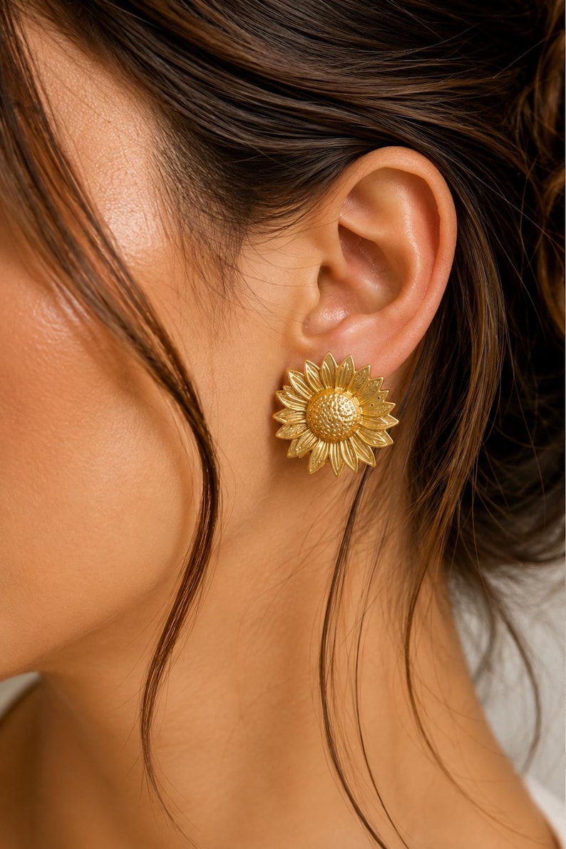 May include: Gold-coloured sunflower earrings. The earrings feature detailed petals and a textured centre, resembling a real sunflower. The earrings are worn on an ear, with the person's skin visible in the background.