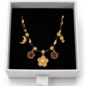 May include: A necklace featuring a black cord with gold and white patterns, adorned with five gold-toned charms including a central flower with a white pearl center, two blue and gold flower charms, a crescent moon, and a small flower charm.