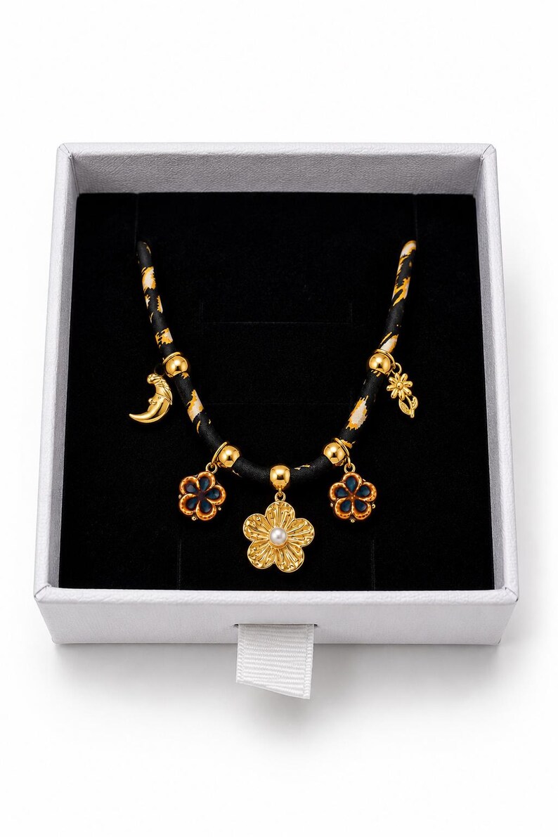 May include: A necklace featuring a black cord with gold and white patterns, adorned with five gold-toned charms including a central flower with a white pearl center, two blue and gold flower charms, a crescent moon, and a small flower charm.