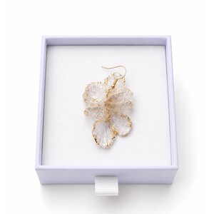 May include: A floral-shaped earring with a gold-colored outline and a clear, petal-like design, presented in a white box with a lavender rim. The earring has a gold-colored hook.