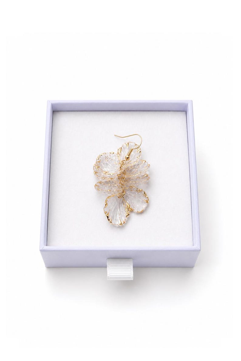 May include: A floral-shaped earring with a gold-colored outline and a clear, petal-like design, presented in a white box with a lavender rim. The earring has a gold-colored hook.