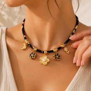 May include: A choker necklace featuring a black and tan animal print fabric band adorned with gold-toned charms, including a crescent moon, two dark blue enamel flowers, a central gold flower with a white pearl center, and a small sun charm.