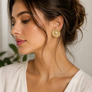 May include: Gold-coloured sunflower stud earrings. The earrings feature textured petals and a textured centre, resembling a real sunflower. The earrings are worn on the ear of a person with dark hair and a white top.