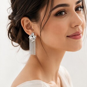 May include: Silver earrings featuring a textured floral design and chain fringe. The earrings are worn by a person with dark hair and a white top. These earrings are a statement piece, adding a touch of elegance to any outfit.