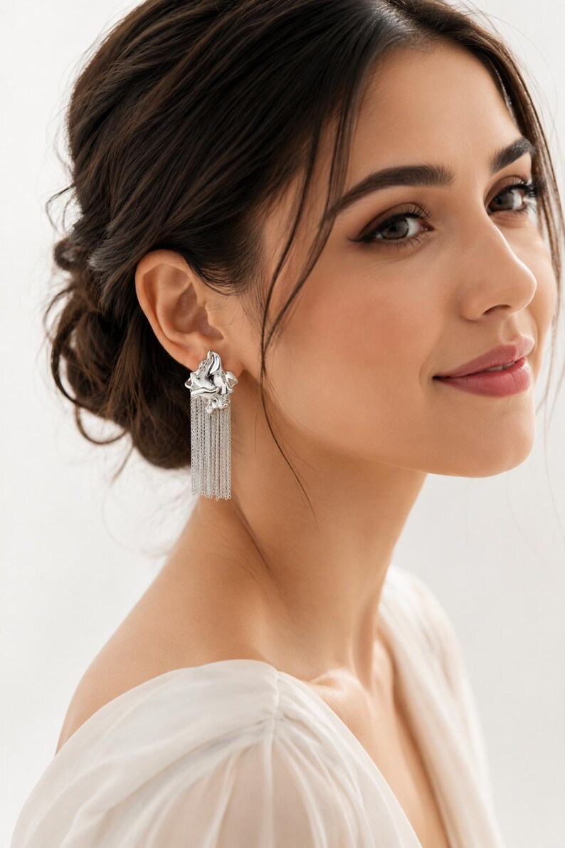 May include: Silver earrings featuring a textured floral design and chain fringe. The earrings are worn by a person with dark hair and a white top. These earrings are a statement piece, adding a touch of elegance to any outfit.