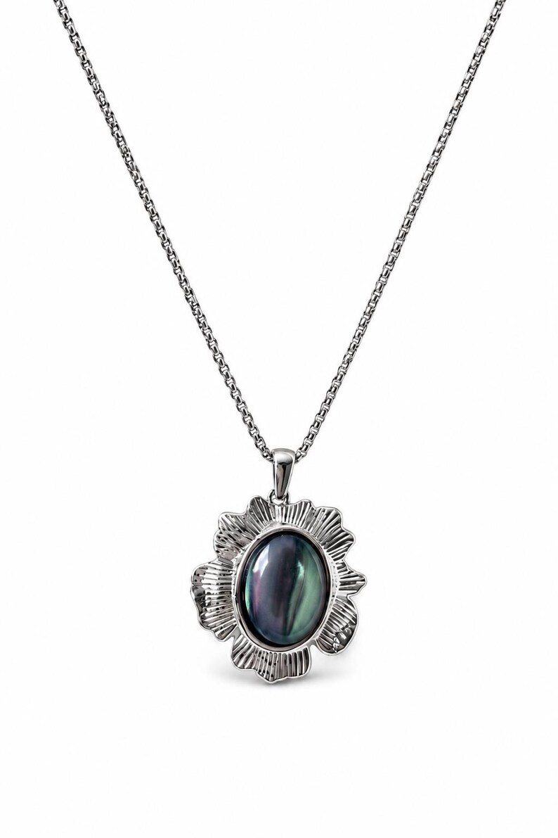 Pode incluir: Um colar de tom prateado com um pendente que apresenta uma pedra escura iridescente de forma oval no centro. A pedra &eacute; rodeada por uma moldura de metal texturizada e recortada que lembra uma flor, suspensa por uma corrente fina prateada.