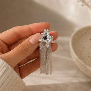 May include: A silver earring with an abstract, star-shaped top and multiple dangling chains. The chains create a fringe effect. The earring is shown against a neutral background. The design is modern and eye-catching, suitable for various occasions.