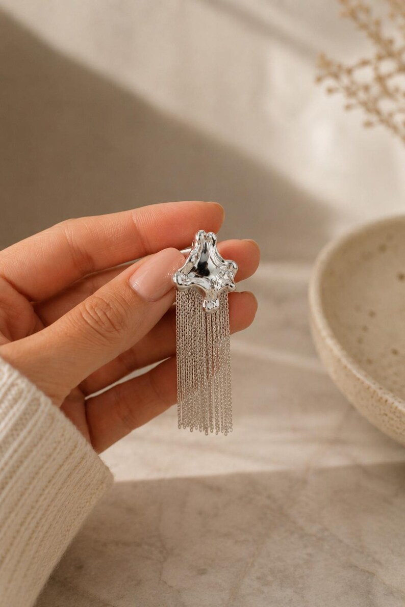 May include: A silver earring with an abstract, star-shaped top and multiple dangling chains. The chains create a fringe effect. The earring is shown against a neutral background. The design is modern and eye-catching, suitable for various occasions.