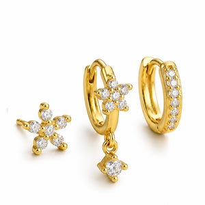 May include: A set of three gold-toned earrings featuring clear, sparkling stones. One earring is a flower-shaped stud, one is a hoop with a flower-shaped charm and a dangling stone, and the third is a hoop with a row of embedded stones.
