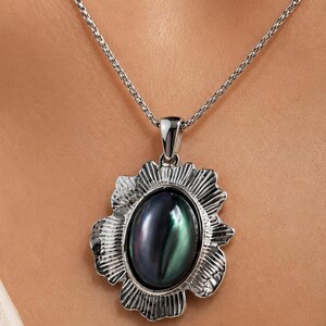 Pode incluir: Um colar com pendente em tom prateado com uma pedra central oval iridescente com reflexos escuros, verdes e roxos. A pedra est&aacute; inserida numa moldura de metal prateado texturizado e recortado com estrias verticais, suspensa numa corrente.