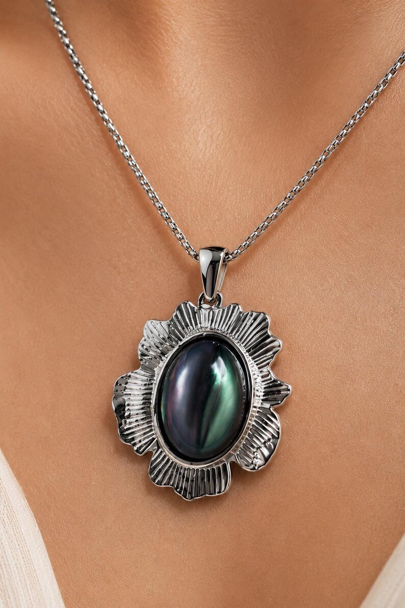 Pode incluir: Um colar com pendente em tom prateado com uma pedra central oval iridescente com reflexos escuros, verdes e roxos. A pedra est&aacute; inserida numa moldura de metal prateado texturizado e recortado com estrias verticais, suspensa numa corrente.