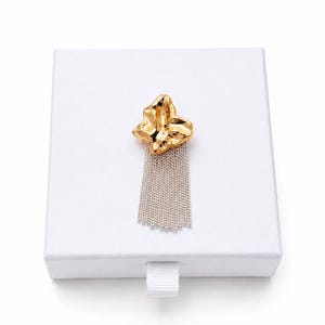 May include: A gold-colored brooch with a textured, abstract design and a silver chain fringe. The brooch is displayed on a white box, suggesting it's a jewelry item. The design is modern and eye-catching.