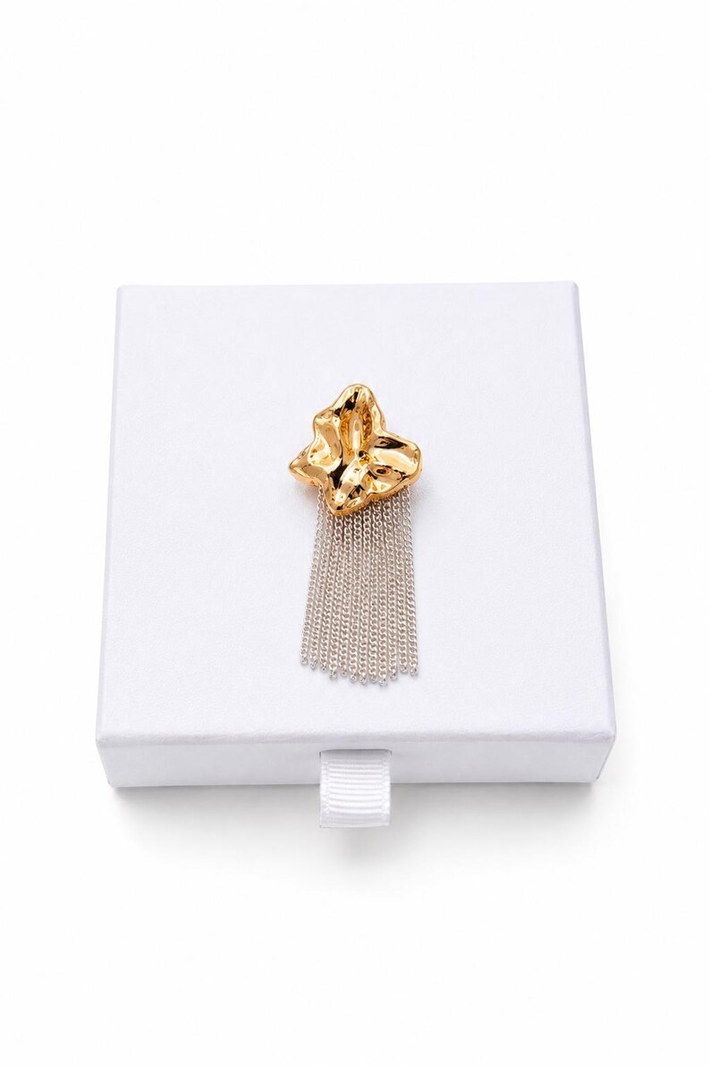 May include: A gold-colored brooch with a textured, abstract design and a silver chain fringe. The brooch is displayed on a white box, suggesting it's a jewelry item. The design is modern and eye-catching.