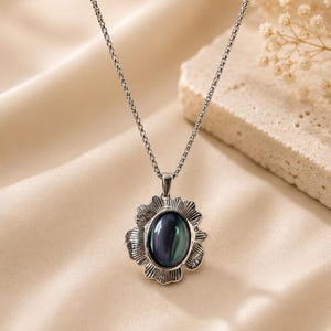 Pode incluir: Um colar de tom prateado com um pendente que apresenta uma pedra central oval de cor escura iridescente. A pedra &eacute; rodeada por uma moldura de metal texturizada e recortada que lembra uma flor. O pendente est&aacute; suspenso numa corrente fina.