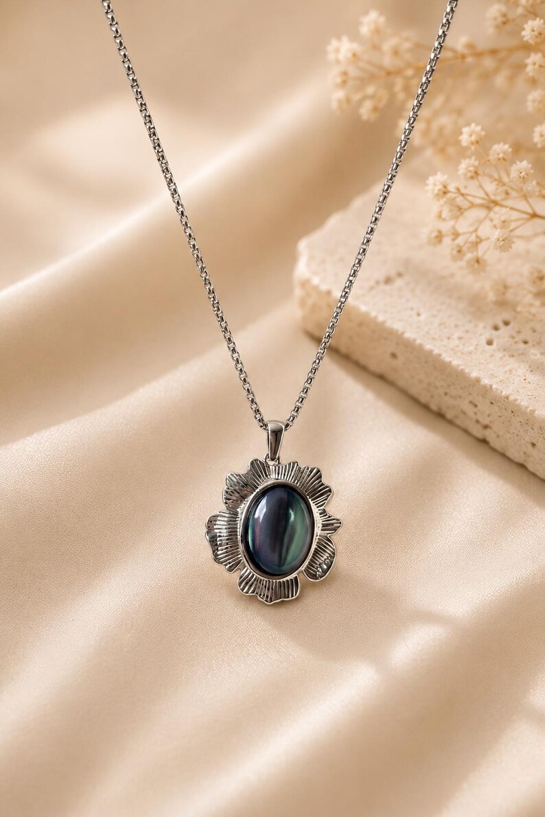 Pode incluir: Um colar de tom prateado com um pendente que apresenta uma pedra central oval de cor escura iridescente. A pedra &eacute; rodeada por uma moldura de metal texturizada e recortada que lembra uma flor. O pendente est&aacute; suspenso numa corrente fina.