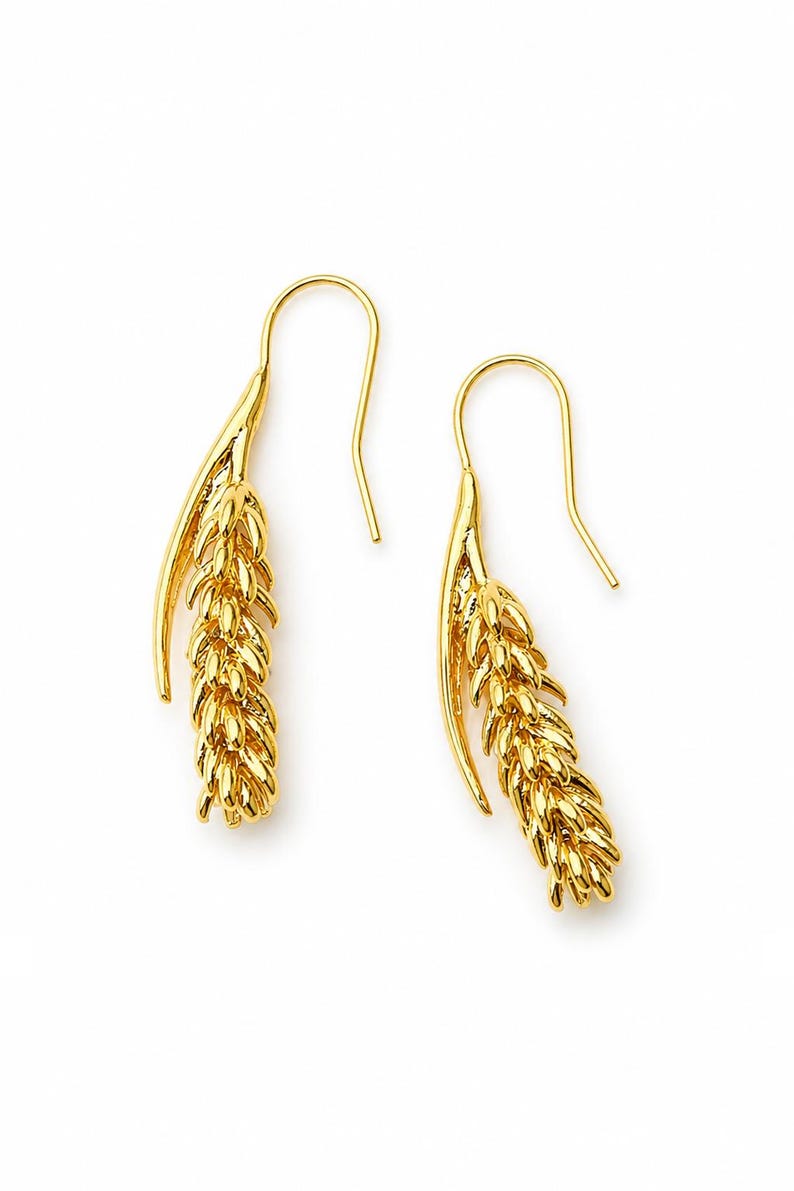May include: A pair of gold-colored earrings. Each earring features a stylized wheat stalk design, with a curved hook for the ear. The earrings are set against a white background, highlighting their intricate details and golden hue.