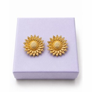 May include: Two gold-coloured sunflower earrings, each with detailed petal and centre designs. The earrings are presented on a lavender-coloured box, creating a striking contrast. The earrings are a jewellery item.