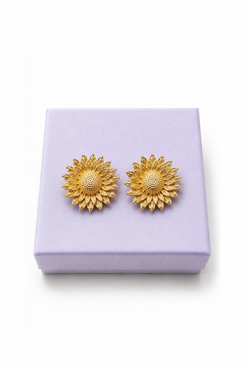 May include: Two gold-coloured sunflower earrings, each with detailed petal and centre designs. The earrings are presented on a lavender-coloured box, creating a striking contrast. The earrings are a jewellery item.