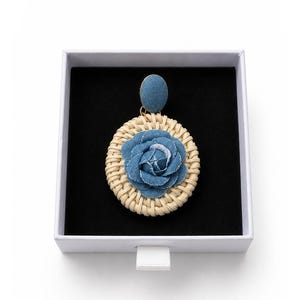May include: A single earring featuring a circular woven rattan base with a blue denim fabric rose at the center. The earring has a blue denim fabric stud top and is presented inside a square white box with a black velvet interior lining.