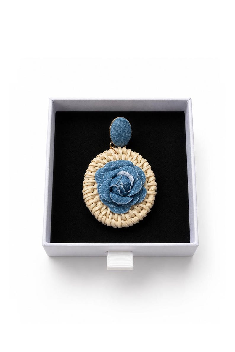 May include: A single earring featuring a circular woven rattan base with a blue denim fabric rose at the center. The earring has a blue denim fabric stud top and is presented inside a square white box with a black velvet interior lining.