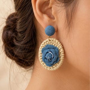 May include: A dangle earring featuring a circular woven rattan frame with a central blue denim fabric rose. The top of the earring consists of a small, round, blue denim-covered stud. The design combines natural beige wicker textures with blue fabric.