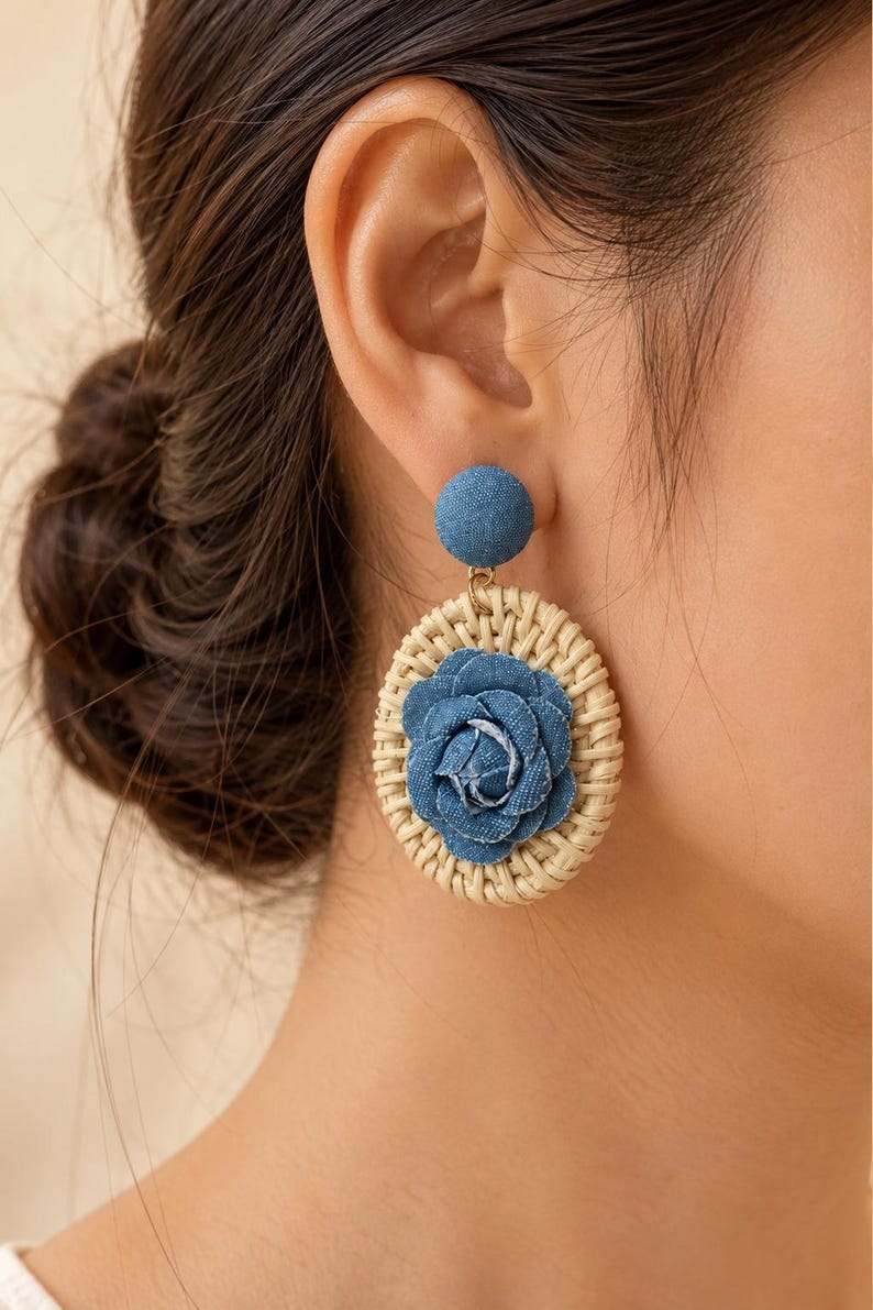 May include: A dangle earring featuring a circular woven rattan frame with a central blue denim fabric rose. The top of the earring consists of a small, round, blue denim-covered stud. The design combines natural beige wicker textures with blue fabric.