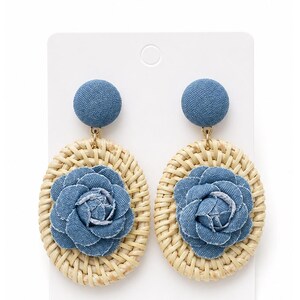 May include: A pair of dangle earrings featuring a circular denim-covered stud top connected to an oval-shaped woven rattan base. Each earring is centered with a layered denim fabric flower, showcasing a blue textile texture and natural tan wicker.