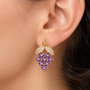 K&ouml;nnte beinhalten: Goldfarbene Ohrringe in Form einer Traube, mit lila Edelsteinen und diamantartigen Akzenten. Die Ohrringe sind an einem Stecker befestigt. Ein Schmuckst&uuml;ck.
