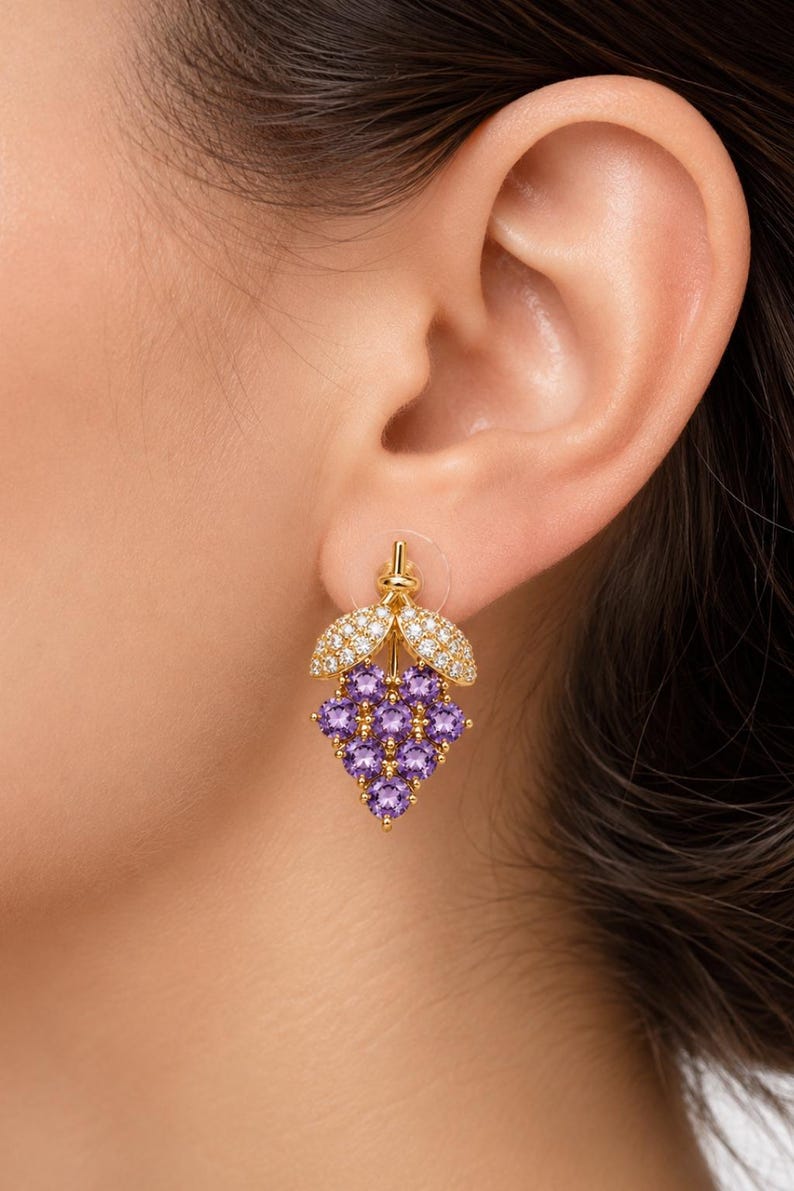K&ouml;nnte beinhalten: Goldfarbene Ohrringe in Form einer Traube, mit lila Edelsteinen und diamantartigen Akzenten. Die Ohrringe sind an einem Stecker befestigt. Ein Schmuckst&uuml;ck.