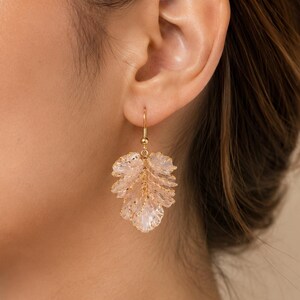 May include: A close-up of a gold-colored earring with a leaf design. The leaf is made of light pink, translucent material with gold accents. The earring is hanging from a gold-colored hook.