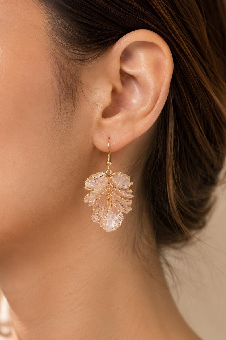 May include: A close-up of a gold-colored earring with a leaf design. The leaf is made of light pink, translucent material with gold accents. The earring is hanging from a gold-colored hook.