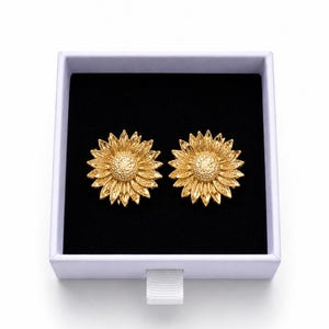 May include: Two gold-coloured sunflower earrings presented in a white box with a black interior. The earrings feature textured petals and a central disc, creating a natural, floral design.