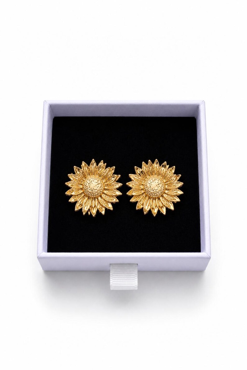 May include: Two gold-coloured sunflower earrings presented in a white box with a black interior. The earrings feature textured petals and a central disc, creating a natural, floral design.