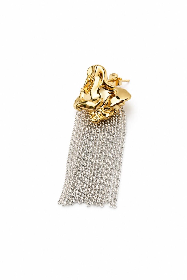 May include: A single gold-colored earring with a textured, organic shape. The earring features a cascade of silver-colored chain fringe, creating a striking contrast. The earring is designed to be worn in a pierced ear.