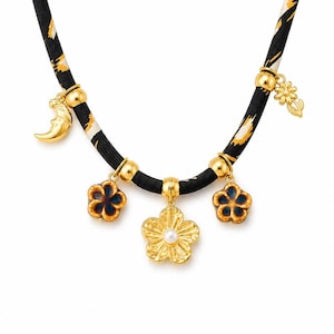 May include: A necklace featuring a black fabric cord with a yellow and white animal print pattern. The cord is adorned with gold-toned metal beads and five charms: a crescent moon, two blue and brown flower charms, a central gold flower with a pearl center, and a small gold flower charm.