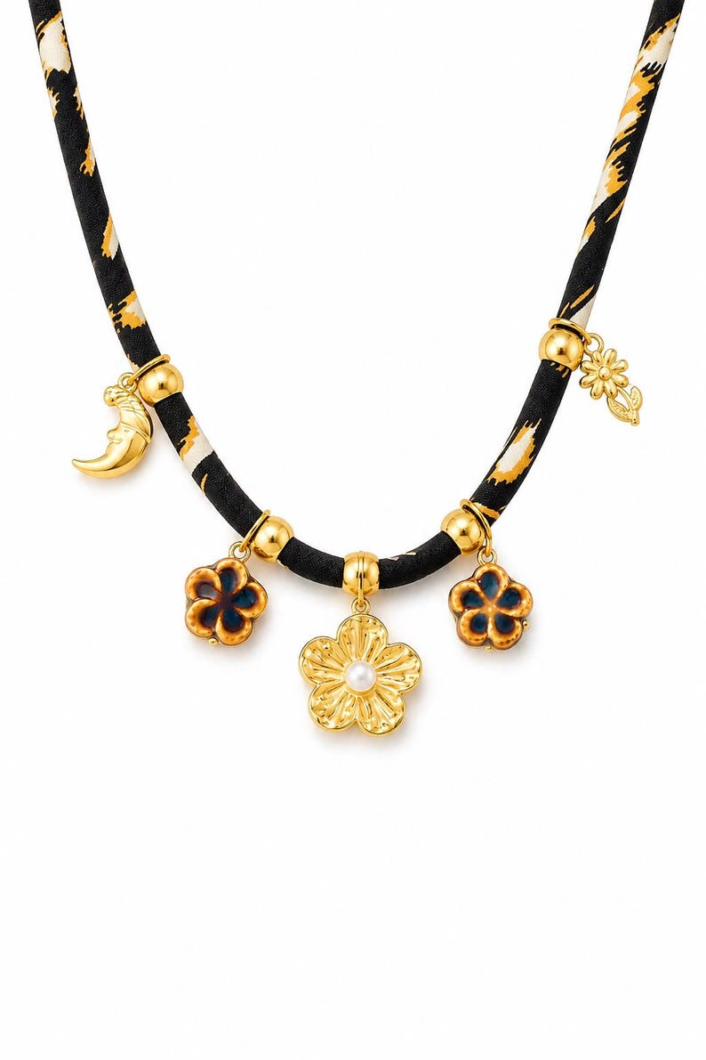 May include: A necklace featuring a black fabric cord with a yellow and white animal print pattern. The cord is adorned with gold-toned metal beads and five charms: a crescent moon, two blue and brown flower charms, a central gold flower with a pearl center, and a small gold flower charm.