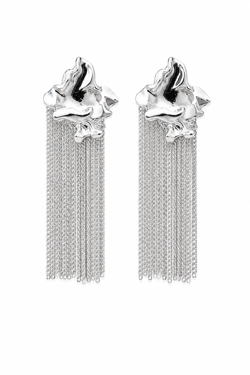 May include: A pair of silver earrings. Each earring has an abstract, textured top with multiple strands of delicate chain hanging down. The earrings are set against a white background, highlighting their reflective surface.