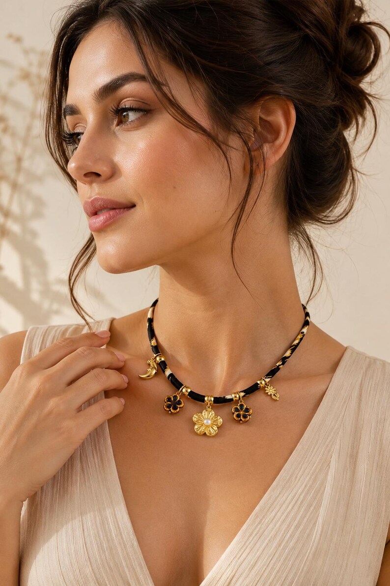 May include: A black and white patterned cord necklace featuring multiple gold-toned charms, including a central flower with a white pearl center, two smaller dark flower charms, and a crescent moon charm, all suspended from gold-toned metal rings.