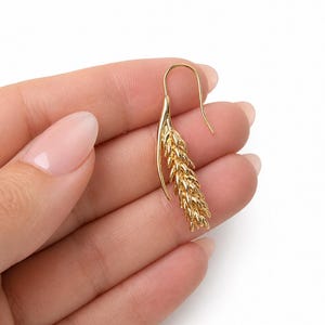 May include: A gold-colored earring with a wheat stalk design. The earring has a curved hook and a detailed, textured wheat stalk pendant. The jewelry is displayed against a white background.