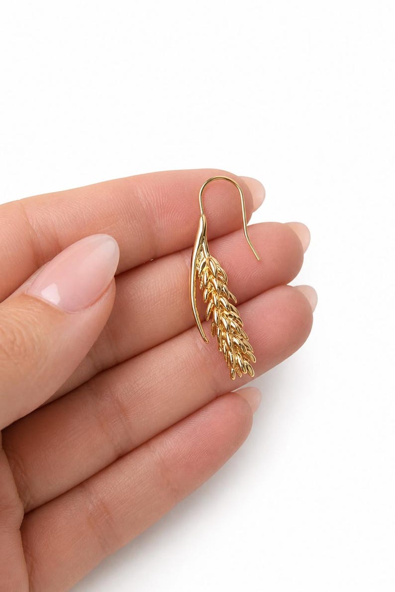 May include: A gold-colored earring with a wheat stalk design. The earring has a curved hook and a detailed, textured wheat stalk pendant. The jewelry is displayed against a white background.