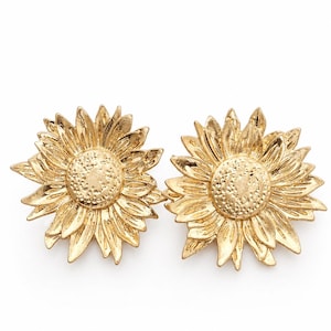 May include: A pair of gold-coloured sunflower earrings. Each earring features a detailed, textured design of a sunflower with petals and a central disc. The earrings are set against a white background.