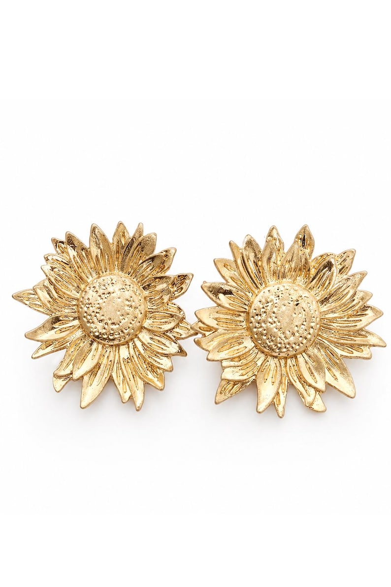 May include: A pair of gold-coloured sunflower earrings. Each earring features a detailed, textured design of a sunflower with petals and a central disc. The earrings are set against a white background.