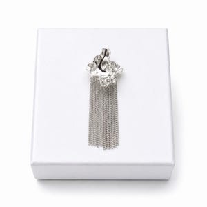May include: A silver brooch with a textured, organic-shaped top and a cascade of dangling chains. The brooch is displayed on a white surface, creating a contrast that highlights its metallic sheen. The design is modern and elegant.